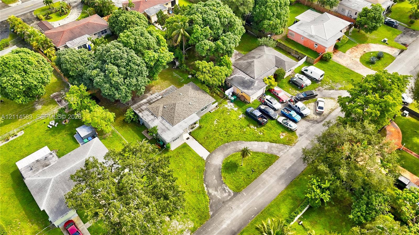 13301 NW Miami Court, Miami, FL 33168 Aerial view of a residential lot featuring a hip roof structure, paved circular driveway, mature perimeter trees, and expansive lawn area