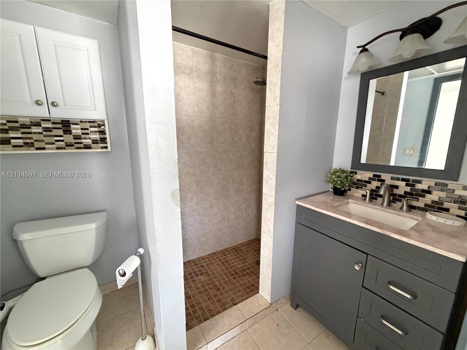 Bathroom featuring a grey vanity with stone-style countertop, integrated sink, tiled shower enclosure, multi-toned decorative backsplash, and wall-mounted vanity lighting