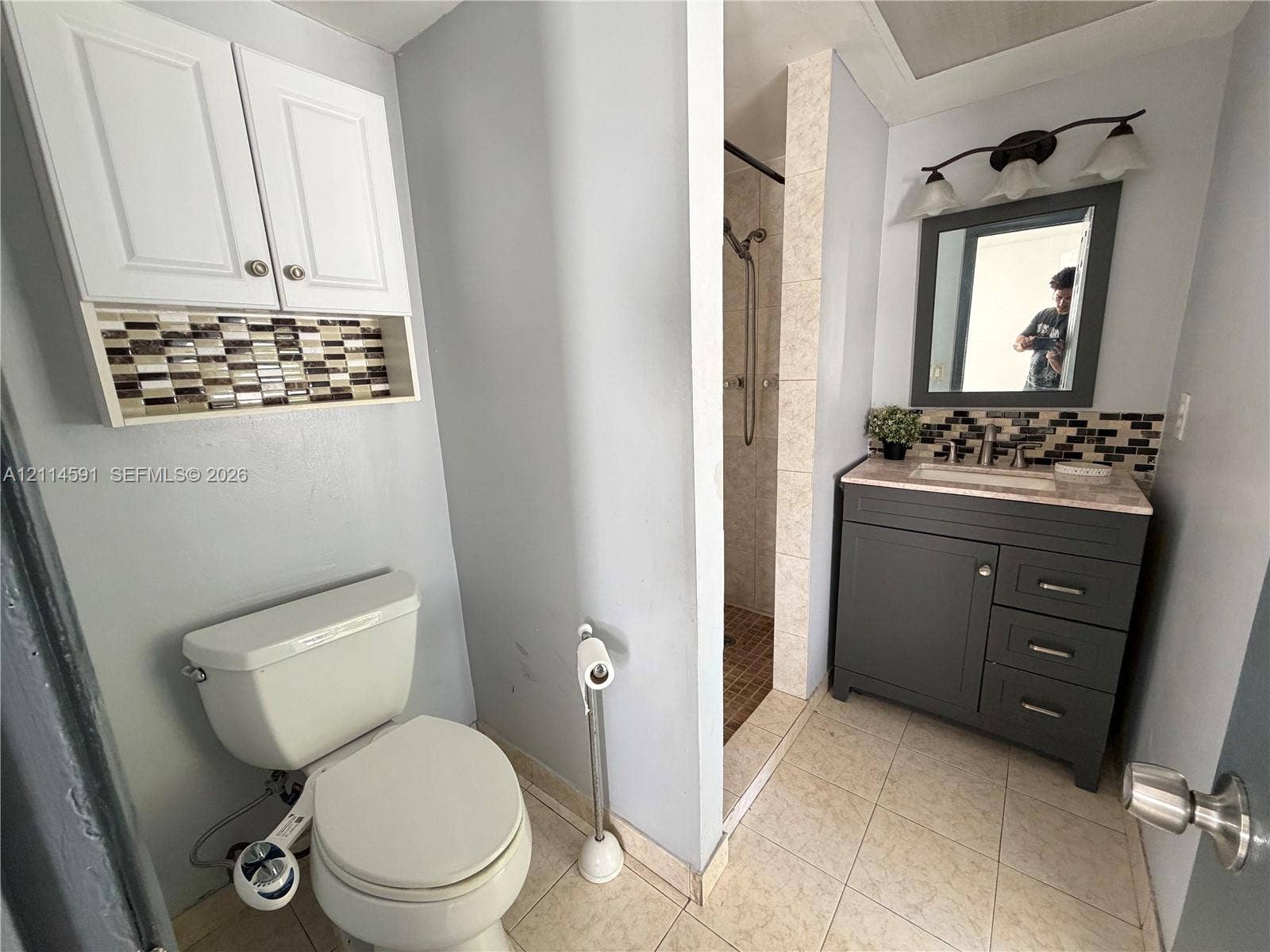 Bathroom featuring a charcoal vanity with a stone countertop, glass mosaic tile backsplash, white wall-mounted upper cabinetry, and a walk-in shower with tiled surround