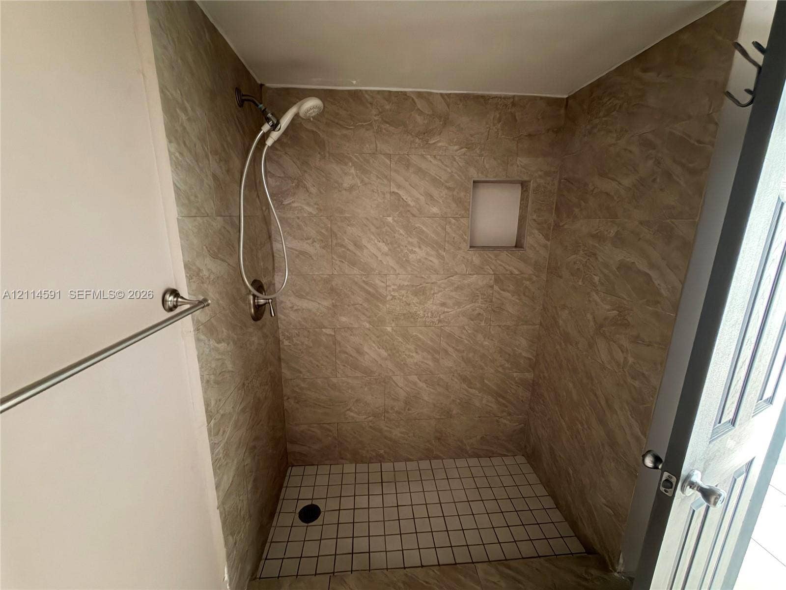 Walk-in shower featuring neutral stone-finish wall tile, a built-in recessed wall niche, a square-tile shower floor, and a polished chrome handheld shower fixture