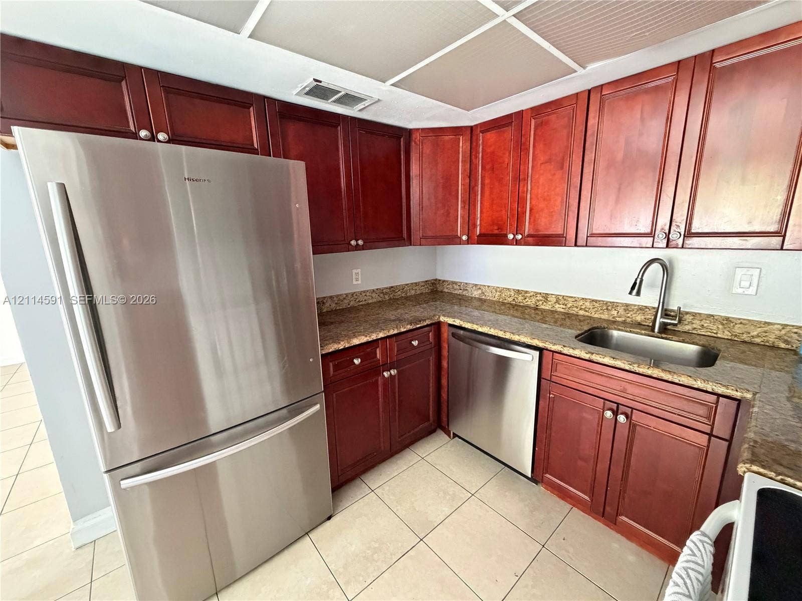 Rich-toned cabinetry, stainless steel refrigerator and dishwasher, stone-finish countertops, stainless steel sink with high-arc faucet, and tiled flooring