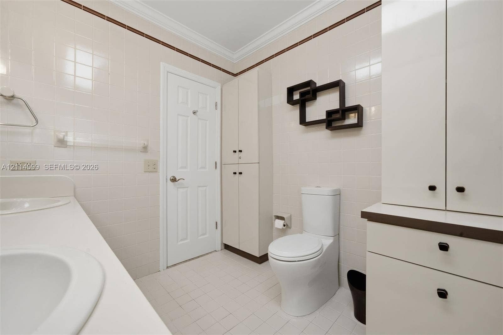 Bathroom featuring floor-to-ceiling ceramic wall tiling, dual-basin vanity with solid surface countertop, crown molding, and extensive built-in cabinetry