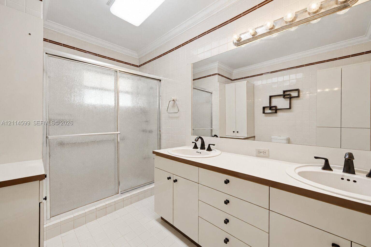 Dual-sink vanity with white countertop, extensive wall-to-wall mirror, shower enclosure with sliding glass doors, crown molding, and tiled wall accents