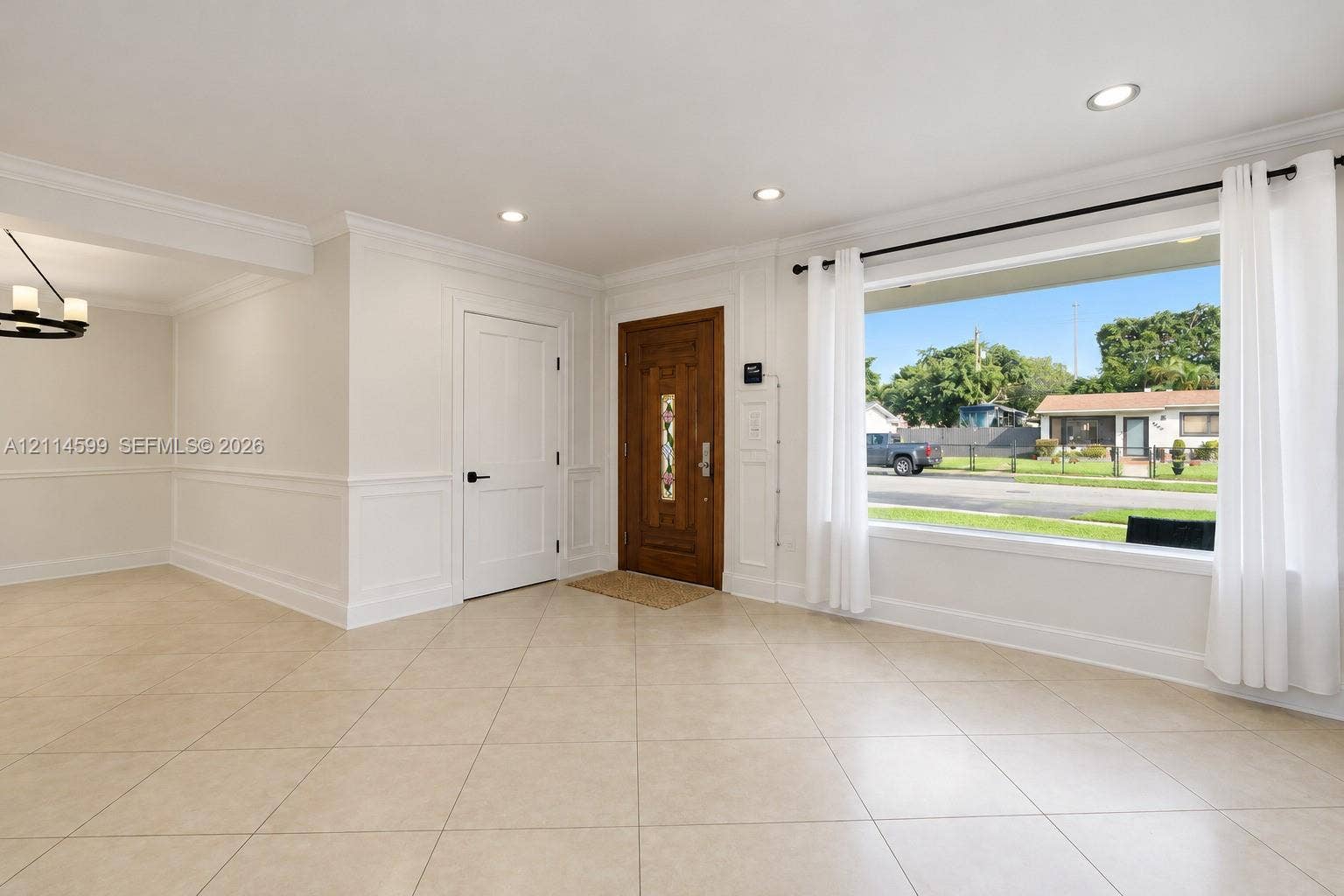 Neutral tile flooring, expansive picture window, wainscoting detail, recessed lighting, and solid wood-finish entry door with decorative glass inlay