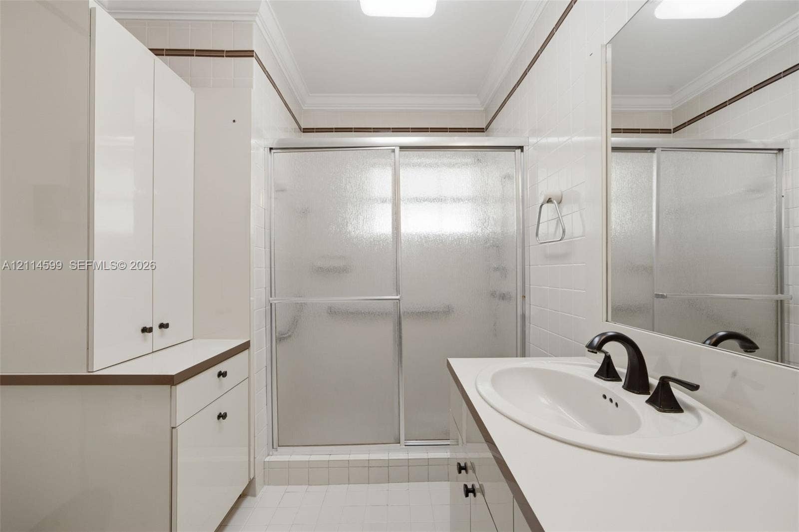 Bathroom featuring white cabinetry, solid-surface countertop, integrated oval sink with dark-finish hardware, glass-enclosed shower, and crown molding