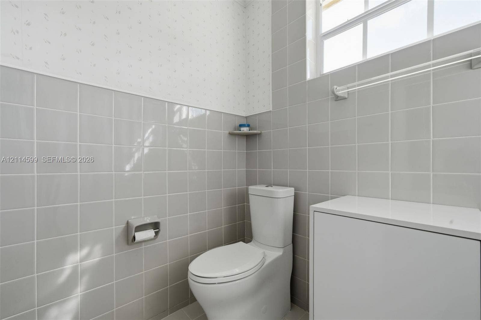 Glossy gray wall tile, dual-flush toilet, white cabinet with solid-surface top, integrated corner shelving, and multi-pane window