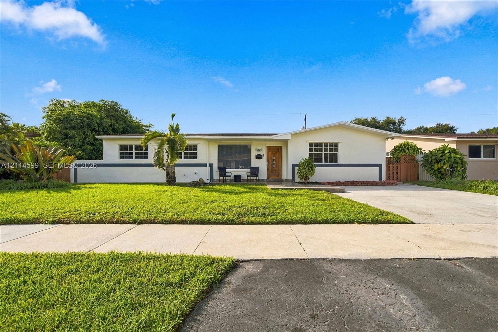 Single-story exterior featuring light-toned stucco siding, horizontal dark-toned accent trim, a concrete driveway, and a front lawn with established landscaping