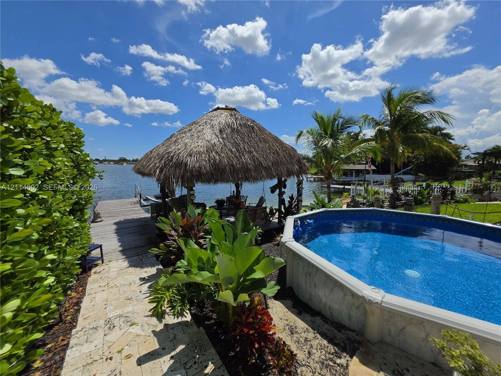 8450 NW 178th Street, Hialeah, Hialeah, FL 33015 Waterfront backyard featuring an above-ground pool, a thatched-roof tiki hut, a wood-plank dock, and a natural stone-paver walkway