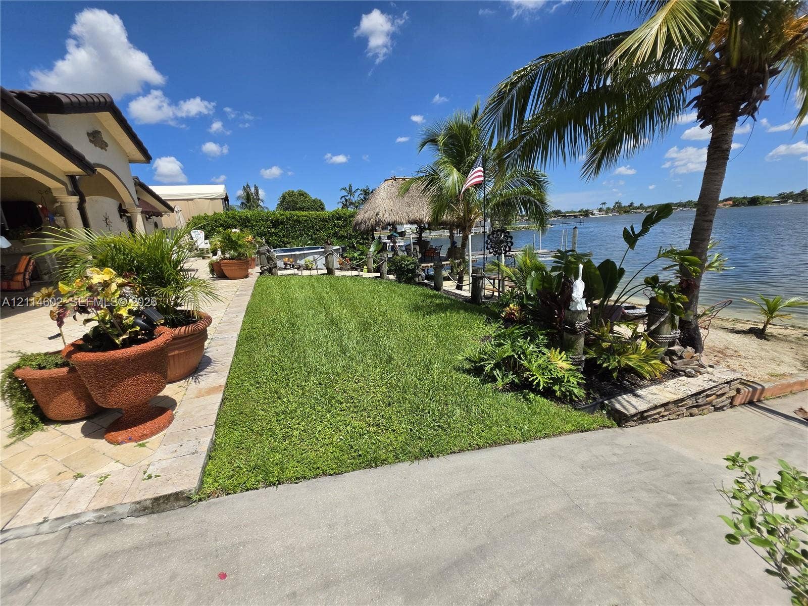 8450 NW 178th Street, Hialeah, Hialeah, FL 33015 Waterfront backyard featuring a manicured lawn, stone-paved patio, mature palm trees, and a thatched-roof structure