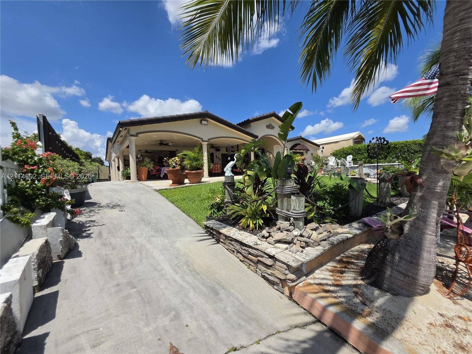 8450 NW 178th Street, Hialeah, Hialeah, FL 33015 Covered patio featuring architectural pillars, concrete driveway, stone-finished retaining wall, and landscaped lawn