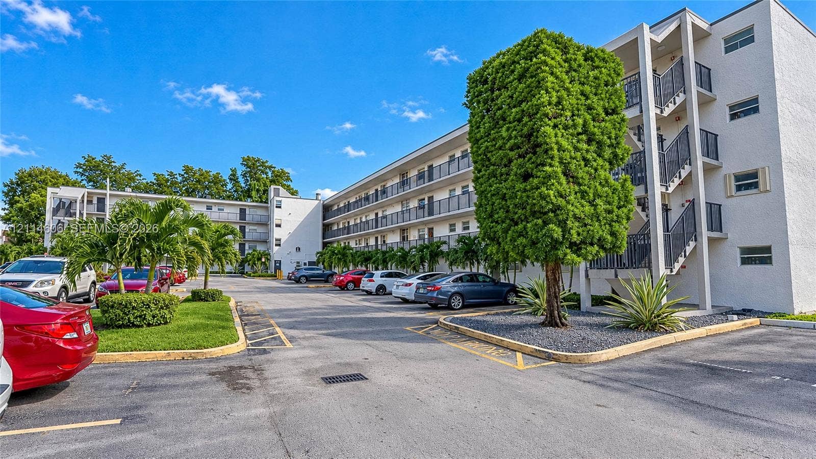 14155 SW 87th Street, #404-E, Miami, FL 33183 Multi-story building exterior featuring a white stucco facade, metal balcony railings, and paved surface parking with landscaped green spaces