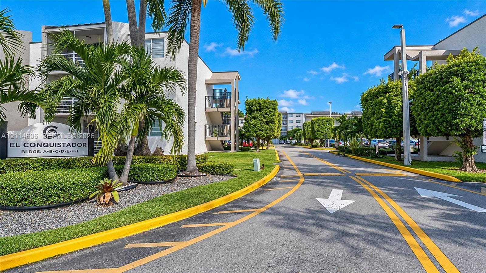 14155 SW 87th Street, #404-E, Miami, FL 33183 Exterior entrance featuring stucco-finished buildings, mature palm trees, manicured hedging, and paved roadway with directional pavement markings