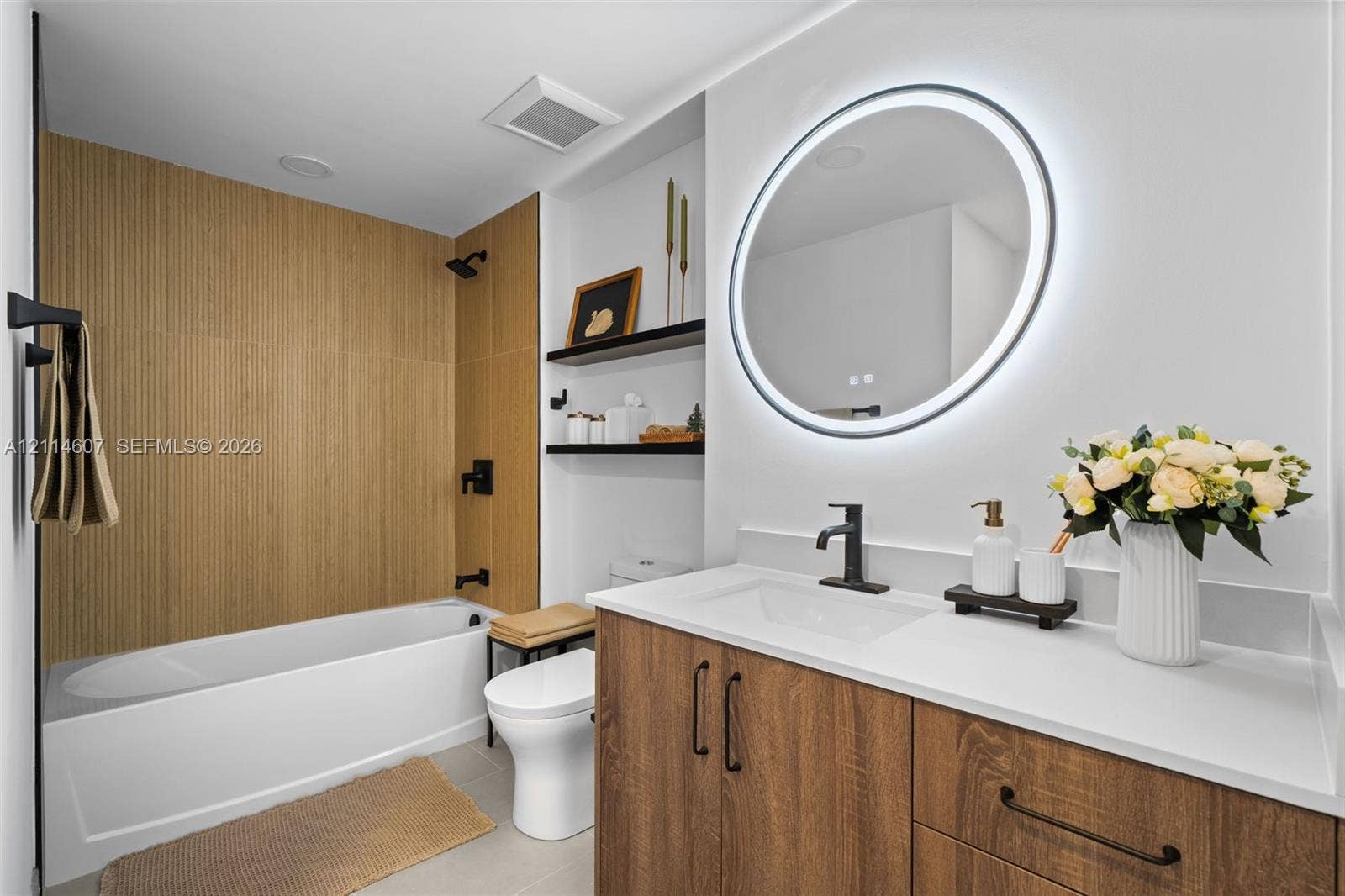 2479 NE 192nd Street, #103, Miami, Miami, FL 33180 Bathroom featuring a vanity with wood-finish cabinetry, quartz countertop, and integrated sink, illuminated round LED mirror, and a tub with wood-finish tile surround and matte black fixtures