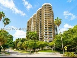 High-rise residential tower featuring curved architectural design, private wrap-around balconies, and extensive exterior glass elements, situated among mature palm trees and landscaped grounds