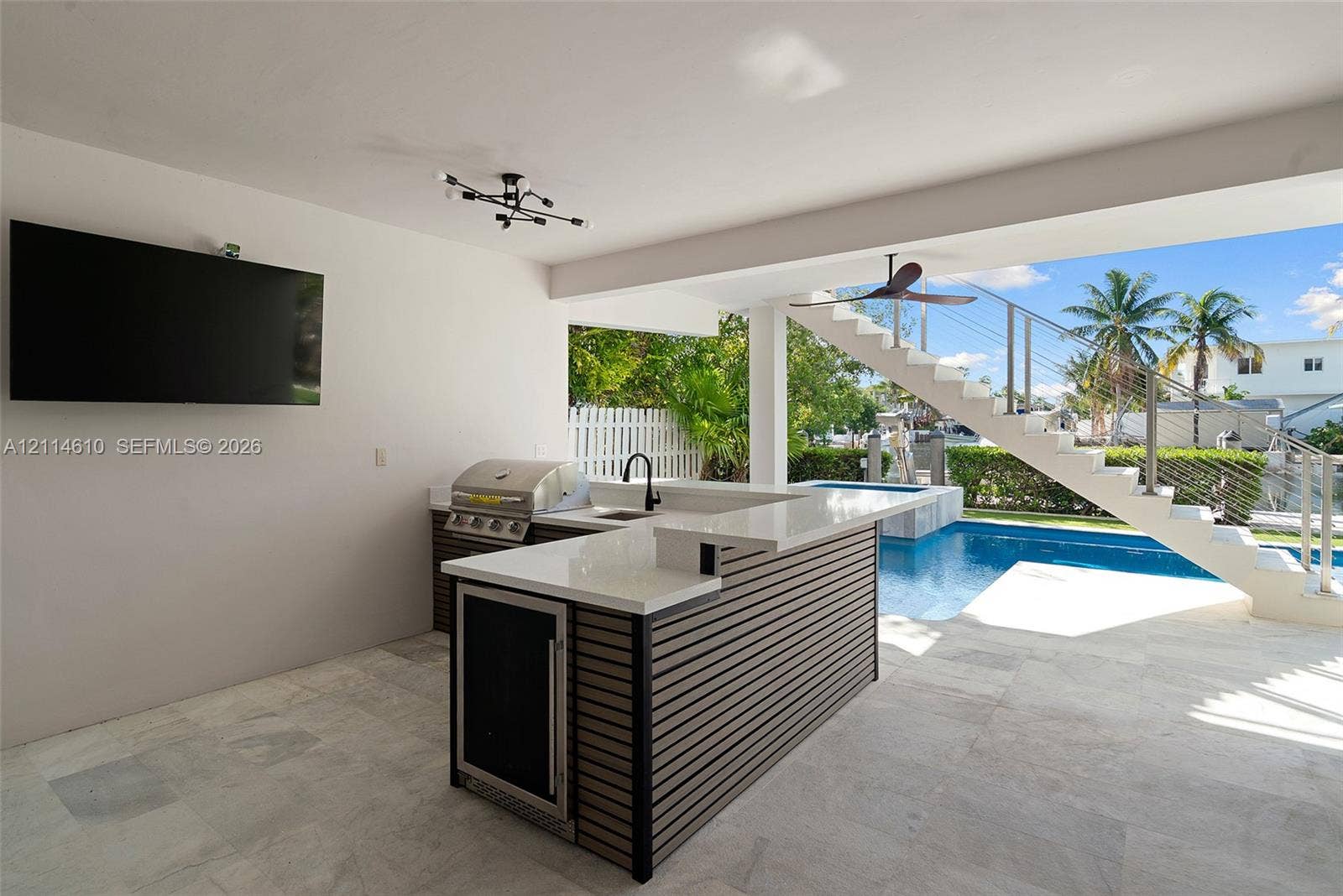 131 Ojibway Avenue, Islamorada, FL 33070 Covered outdoor kitchen featuring a stone-topped island with built-in stainless steel appliances, sleek slatted base cabinetry, stone tile flooring, and integrated ceiling lighting, situated poolside with access to a concrete staircase