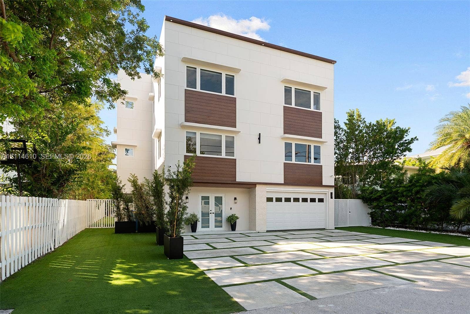 131 Ojibway Avenue, Islamorada, FL 33070 Modern three-story exterior featuring smooth white paneling, horizontal wood-finish siding, a multi-paneled garage door, and a geometric concrete paver driveway with turf accents