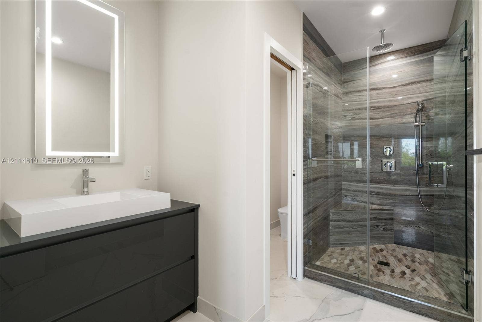 131 Ojibway Avenue, Islamorada, FL 33070 Modern bathroom featuring a dark floating vanity with an integrated rectangular sink, wall-mounted LED-backlit mirror, and a walk-in shower with floor-to-ceiling stone-patterned tile, a rainfall showerhead, and a glass enclosure