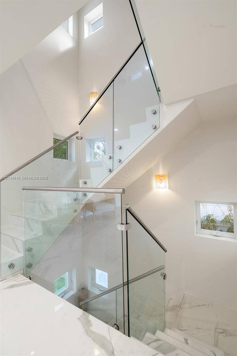131 Ojibway Avenue, Islamorada, FL 33070 Staircase featuring white stone treads, frameless glass railings with metal standoffs, stainless steel handrails, multiple square windows, and wall-mounted light fixtures
