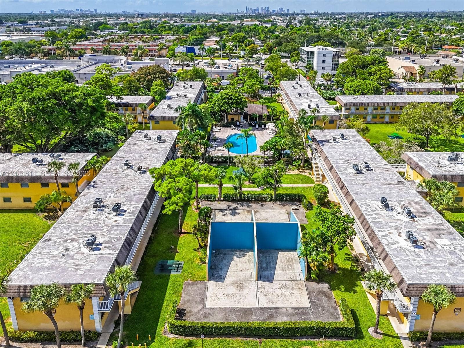 Aerial perspective of a multi-building property featuring a central outdoor swimming pool, dedicated racquetball courts, extensive manicured lawn areas, and mature palm trees