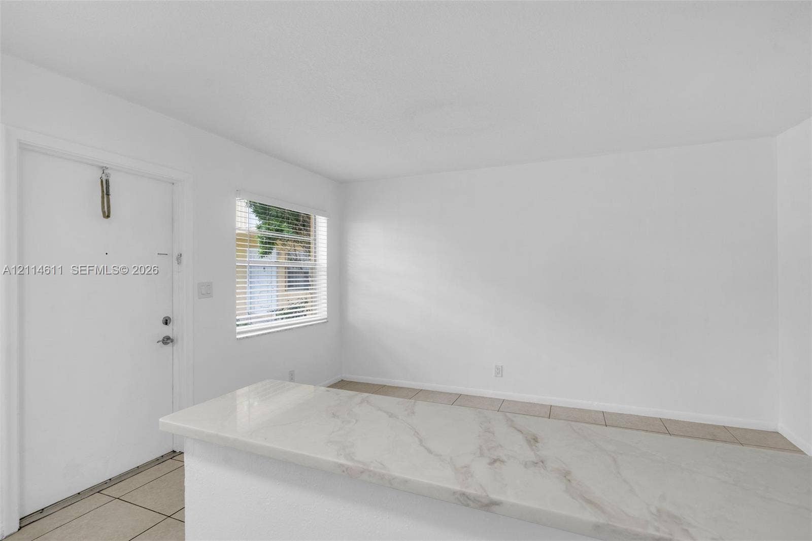 Interior space featuring white walls, neutral tile flooring, horizontal window blinds, and a countertop with a marble-patterned finish