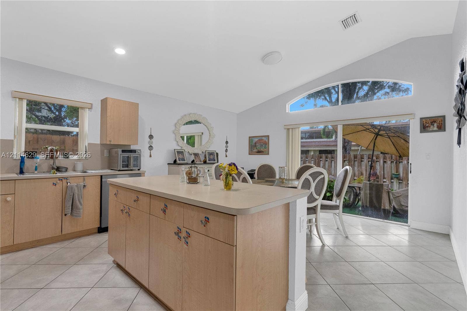 Kitchen featuring a central island with a solid surface countertop, light wood cabinetry, ceramic tile flooring, a vaulted ceiling, and an arched transom window above the sliding glass door