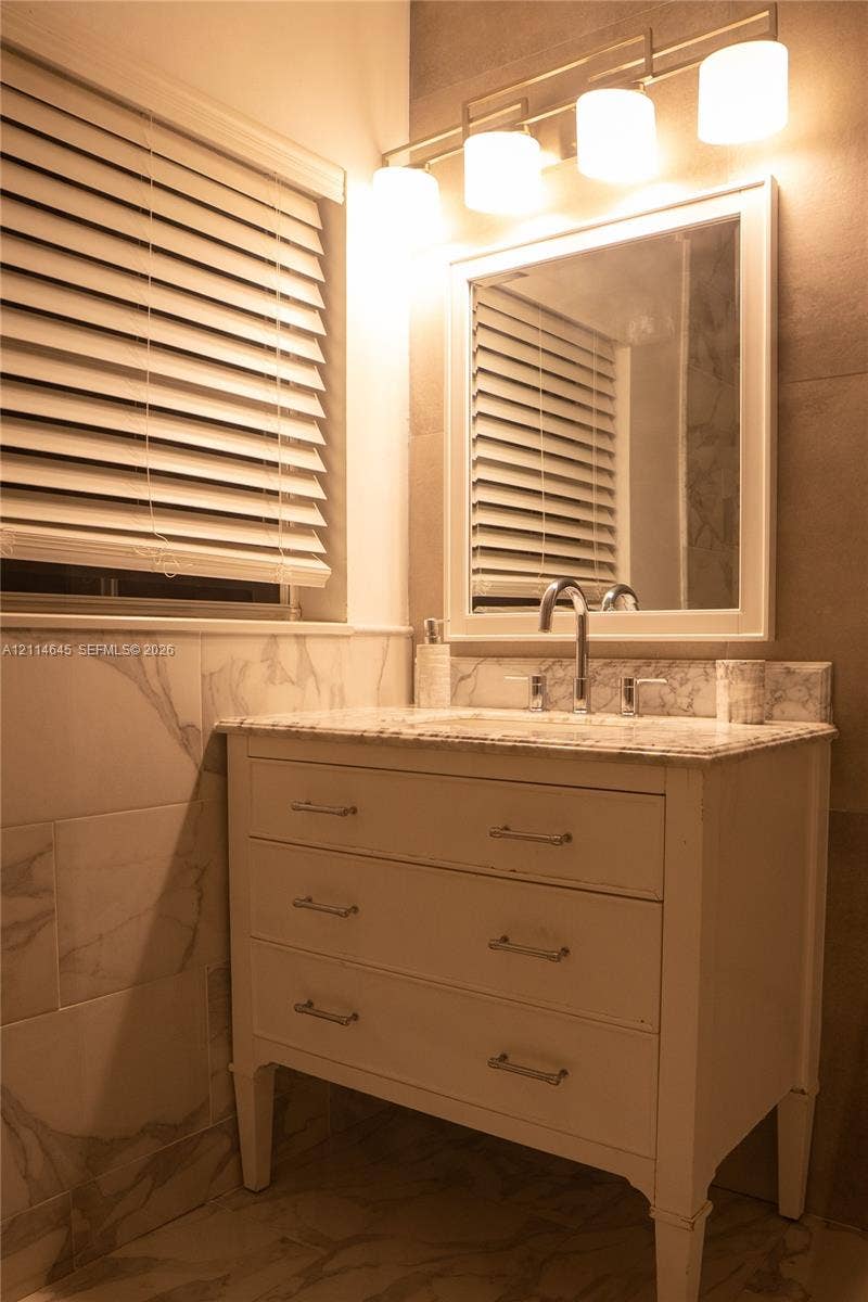 Bathroom vanity featuring a stone countertop, white cabinetry with polished chrome pulls, a framed wall mirror, a four-light fixture, and large-format wall tile
