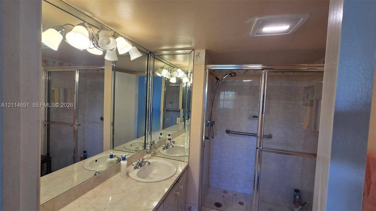 Bathroom vanity featuring dual inset sinks, stone-finish countertops, wall-to-wall mirror, and overhead multi-light fixture, accompanied by a glass-enclosed shower with tiled surround