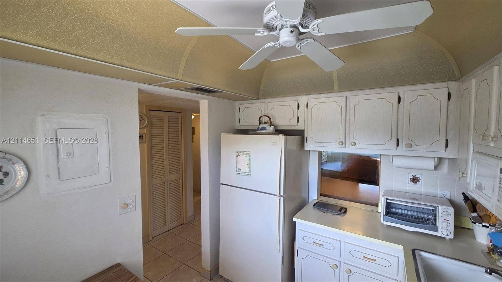 Kitchen featuring white cabinetry with raised-panel doors, neutral solid-surface countertops, tile flooring, a built-in ceiling fan, and curved architectural ceiling detailing
