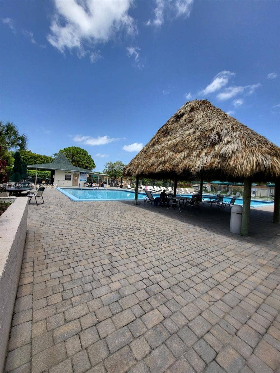 Outdoor recreation area featuring a swimming pool, thatched-roof tiki structure, paved stone deck, and a clubhouse facility