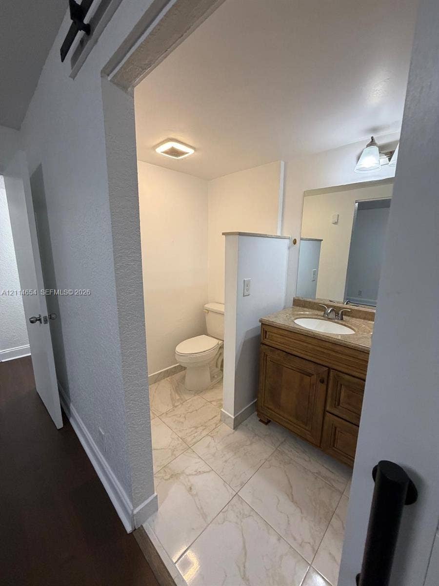 Bathroom featuring large-format stone-finish tile flooring, wood-finish cabinetry, an integrated vanity countertop with an oval basin, and a wall-mounted vanity mirror with a single-light fixture