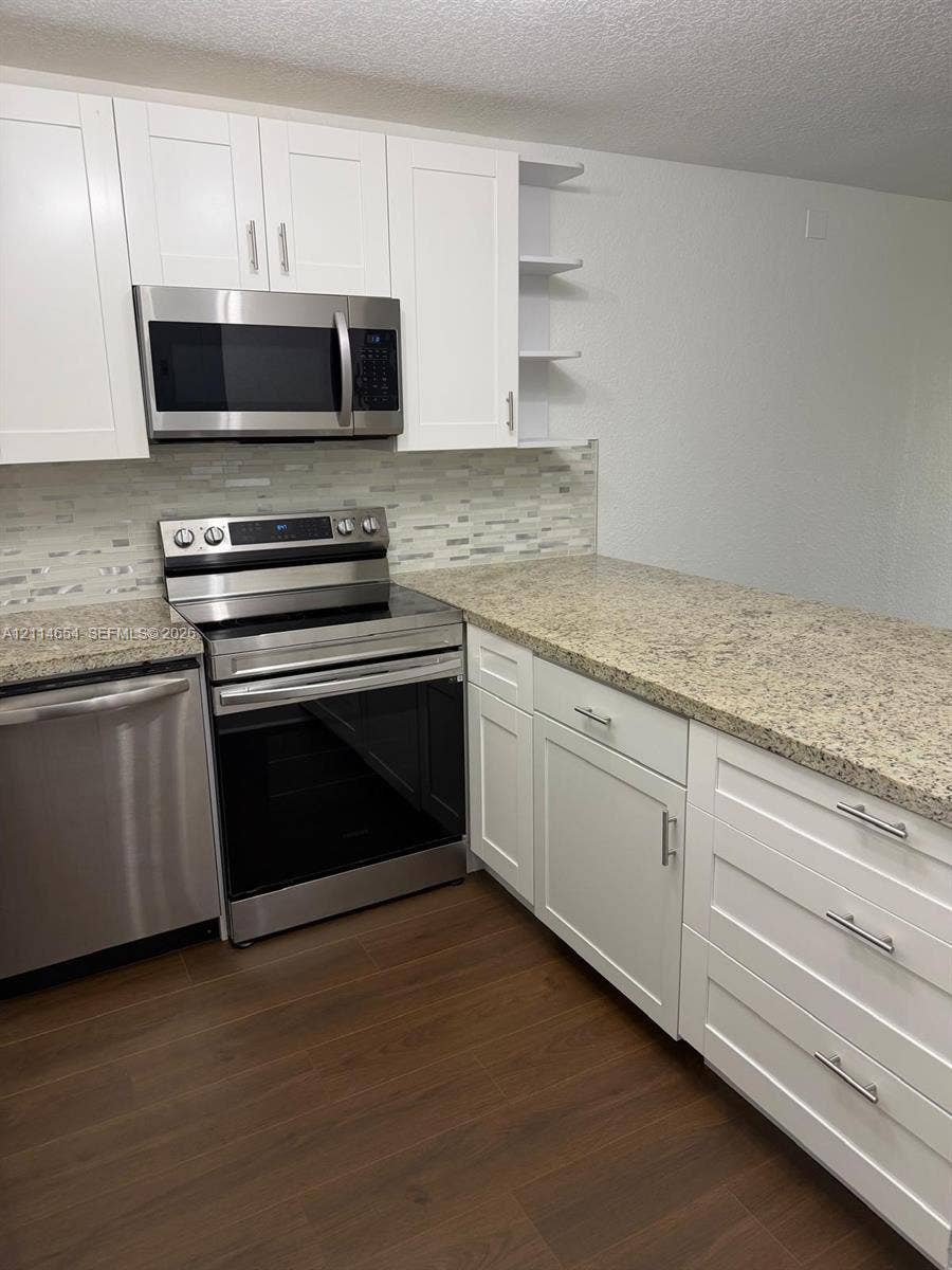 Kitchen featuring white shaker-style cabinetry, stone-finish countertops, stainless steel appliances, a multi-tone tile backsplash, and wood-finish flooring