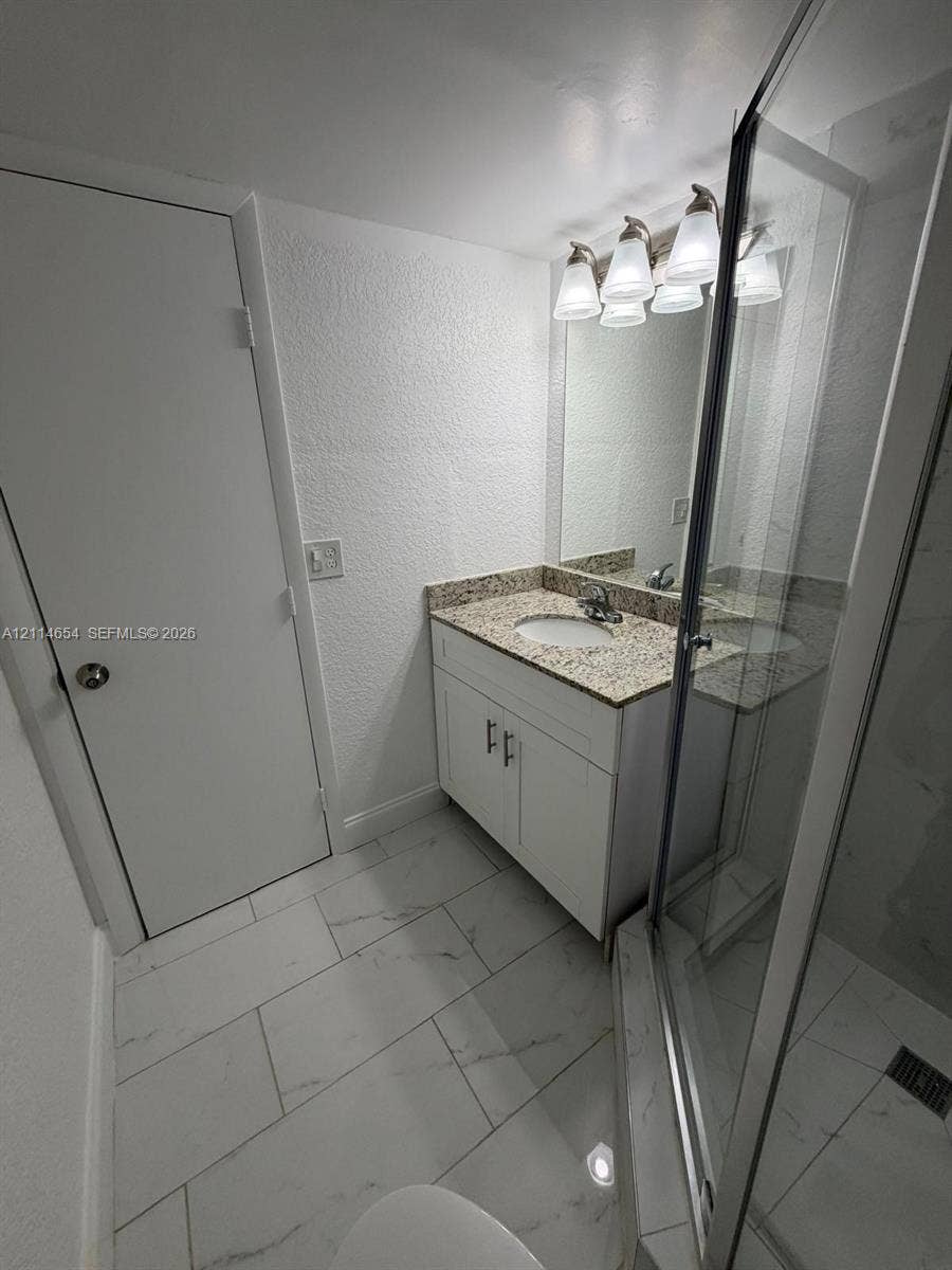 Bathroom featuring white cabinetry, stone-finish countertop with integrated oval sink, glass-enclosed shower, polished light-colored tile flooring, and multi-bulb vanity lighting
