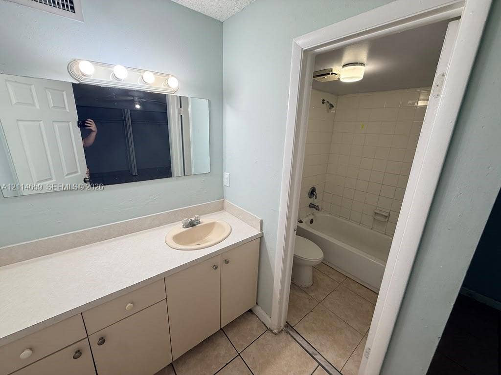 Vanity with integrated basin, solid-surface countertop, cabinetry storage, wall-mounted light fixture, and tiled flooring