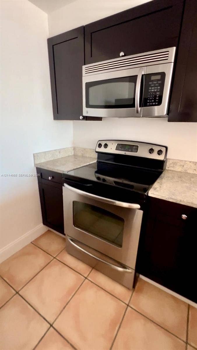 Kitchen featuring dark wood-finish cabinetry, stainless steel microwave, stainless steel electric range, stone-patterned countertops, and ceramic tile flooring