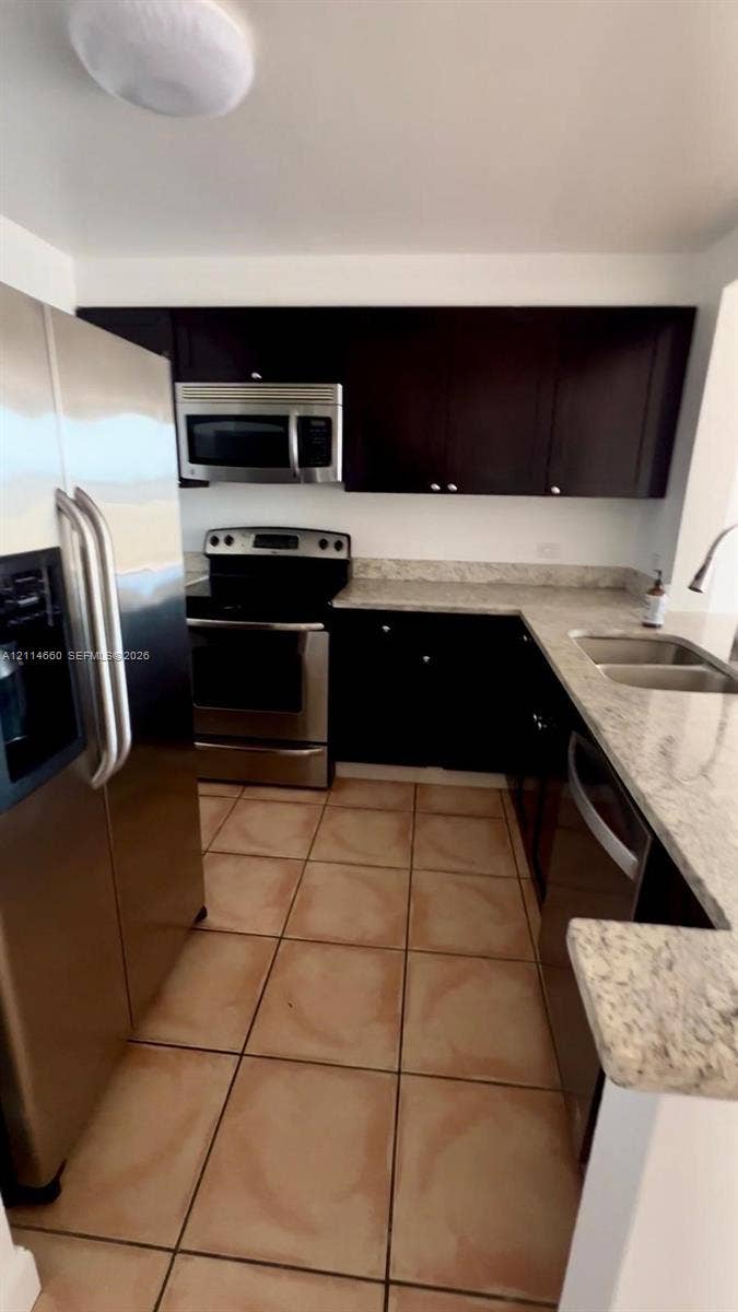 Kitchen featuring dark-finish cabinetry, stone-style countertops, ceramic tile flooring, and stainless steel appliances including a built-in microwave, range, and double-door refrigerator