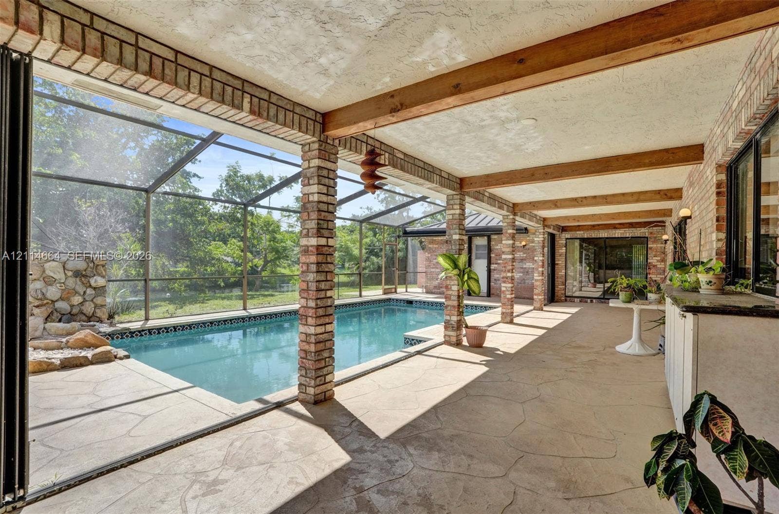 11164 NW 10th Place, Coral Springs, FL 33071 Covered patio featuring exposed wooden beams, brick support columns, a screened-in enclosure, and a swimming pool with a stone rock feature