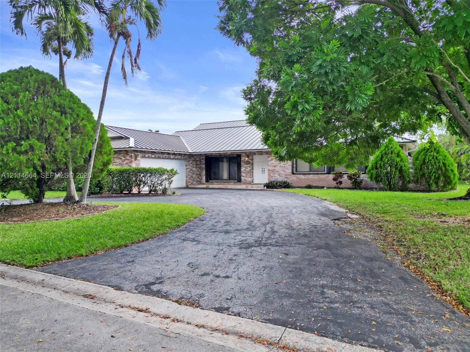 11164 NW 10th Place, Coral Springs, FL 33071 Single-story brick exterior featuring a multi-pitched metal roof, attached garage, and paved driveway surrounded by mature landscaping and established trees
