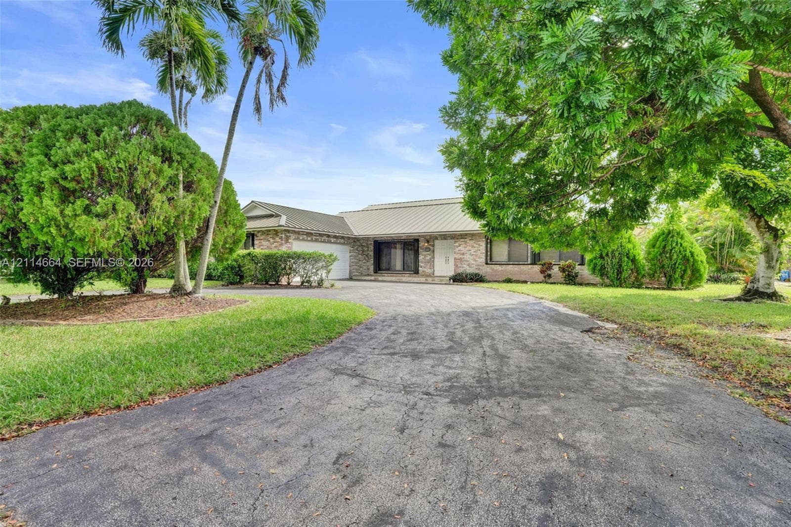 11164 NW 10th Place, Coral Springs, FL 33071 Single-story residence featuring a brick facade, multi-gabled metal roof, attached garage, and mature palm trees situated on a landscaped lot