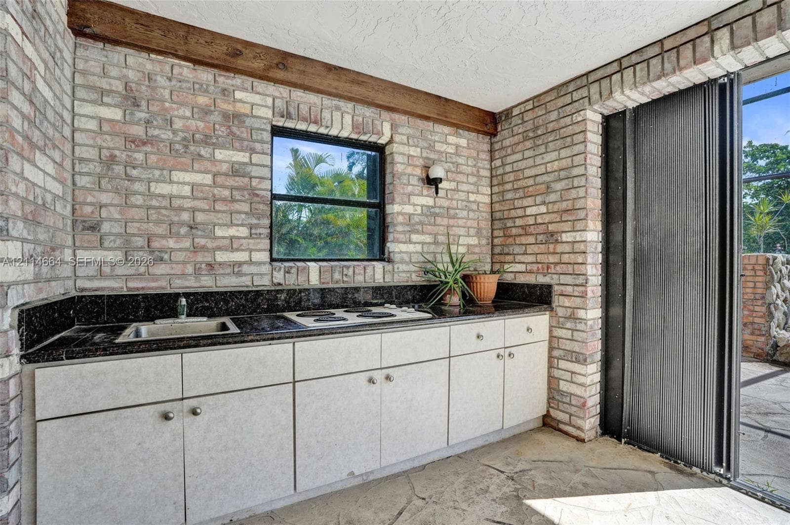 11164 NW 10th Place, Coral Springs, FL 33071 Outdoor kitchen area featuring exposed brick walls, stone flooring, built-in cabinetry, dark stone-finish countertops, and an integrated cooktop and sink