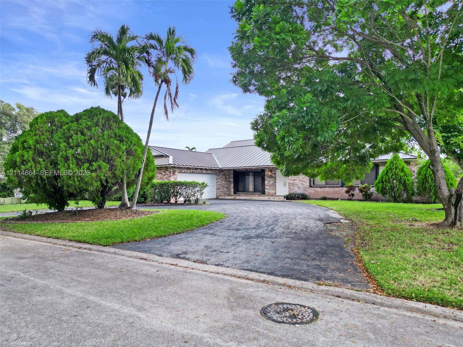 11164 NW 10th Place, Coral Springs, FL 33071 Single-story brick exterior featuring a metal roof, attached two-car garage, mature landscaping, and an expansive paved driveway