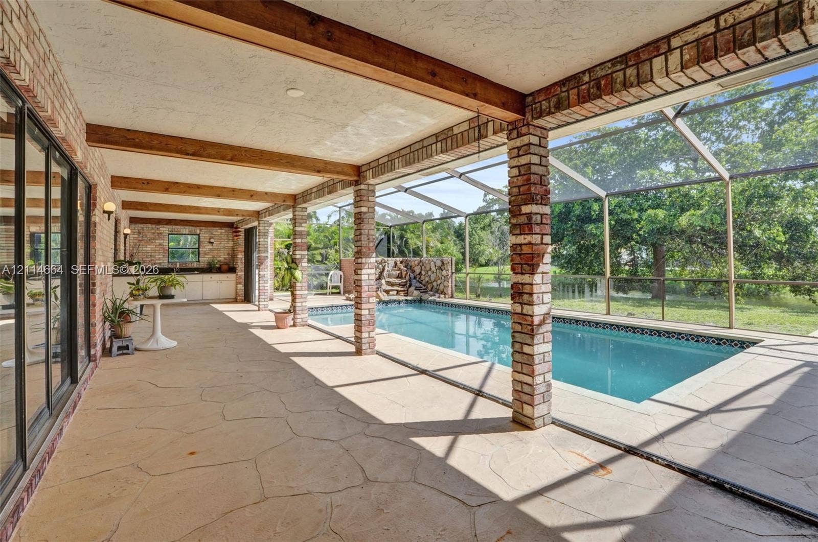 11164 NW 10th Place, Coral Springs, FL 33071 Covered lanai with flagstone flooring, exposed wood ceiling beams, brick support columns, and an in-ground pool enclosed by a screen structure