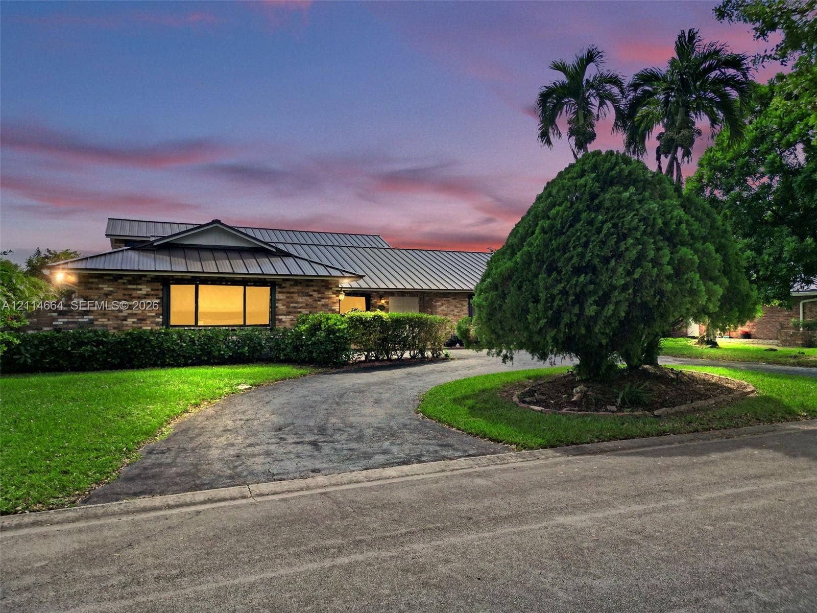 11164 NW 10th Place, Coral Springs, FL 33071 Single-story residence featuring a multi-pitched metal roof, stone veneer exterior, expansive front lawn, circular asphalt driveway, and mature landscaping