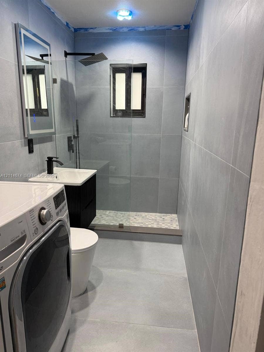 423 NW 32nd Place, Miami, Miami, FL 33125 Walk-in shower with a glass panel, stone-finish shower floor, matte black rainfall showerhead, wall-mounted vanity with a rectangular sink, and illuminated wall mirror