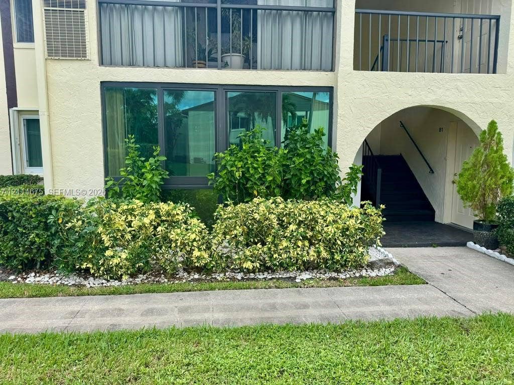 Stucco exterior featuring floor-to-ceiling tinted glass windows, a covered arched entryway, a second-story balcony with metal railings, and landscaped shrubbery