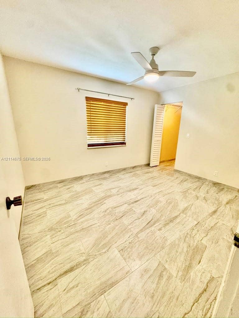 Neutral-toned interior featuring large-format stone-patterned tile flooring, a centered ceiling fan with integrated light fixture, window with horizontal blinds, and a louvered bi-fold closet door