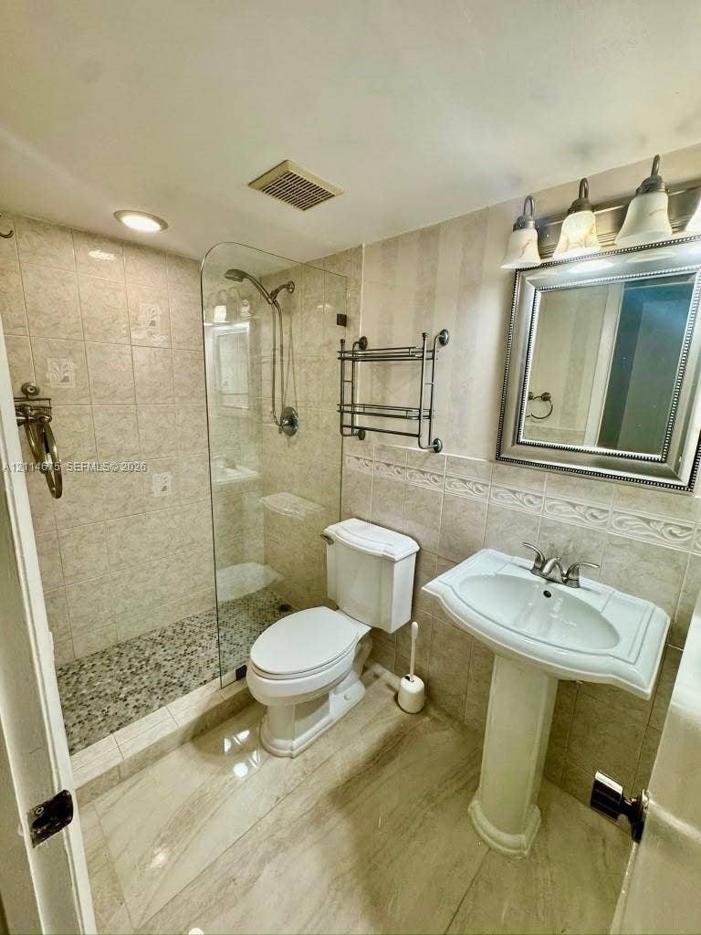 Walk-in shower with a glass panel, stone-finish flooring, pebble-tile shower floor, pedestal sink, and wall-mounted three-light fixture