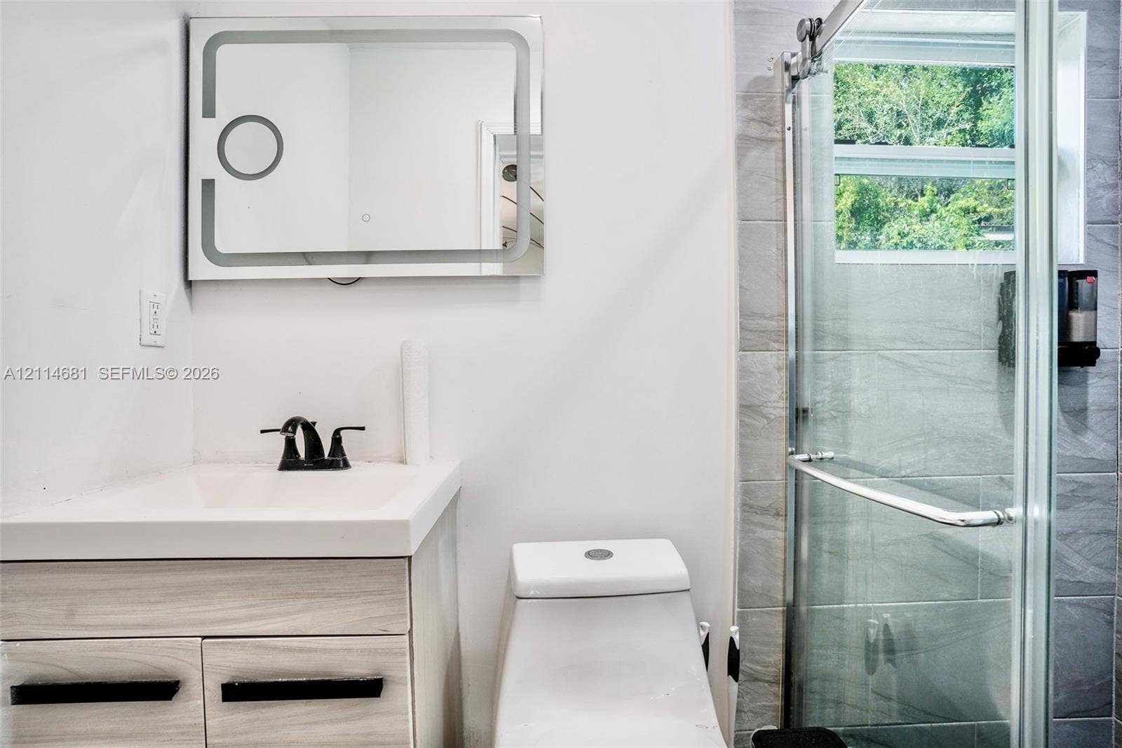 6700 NW 5th Avenue, Miami, FL 33150 Vanity with a white integrated sink, wood-finish cabinetry, matte black hardware, backlit mirror, and a walk-in shower featuring grey tile surround and a sliding glass door