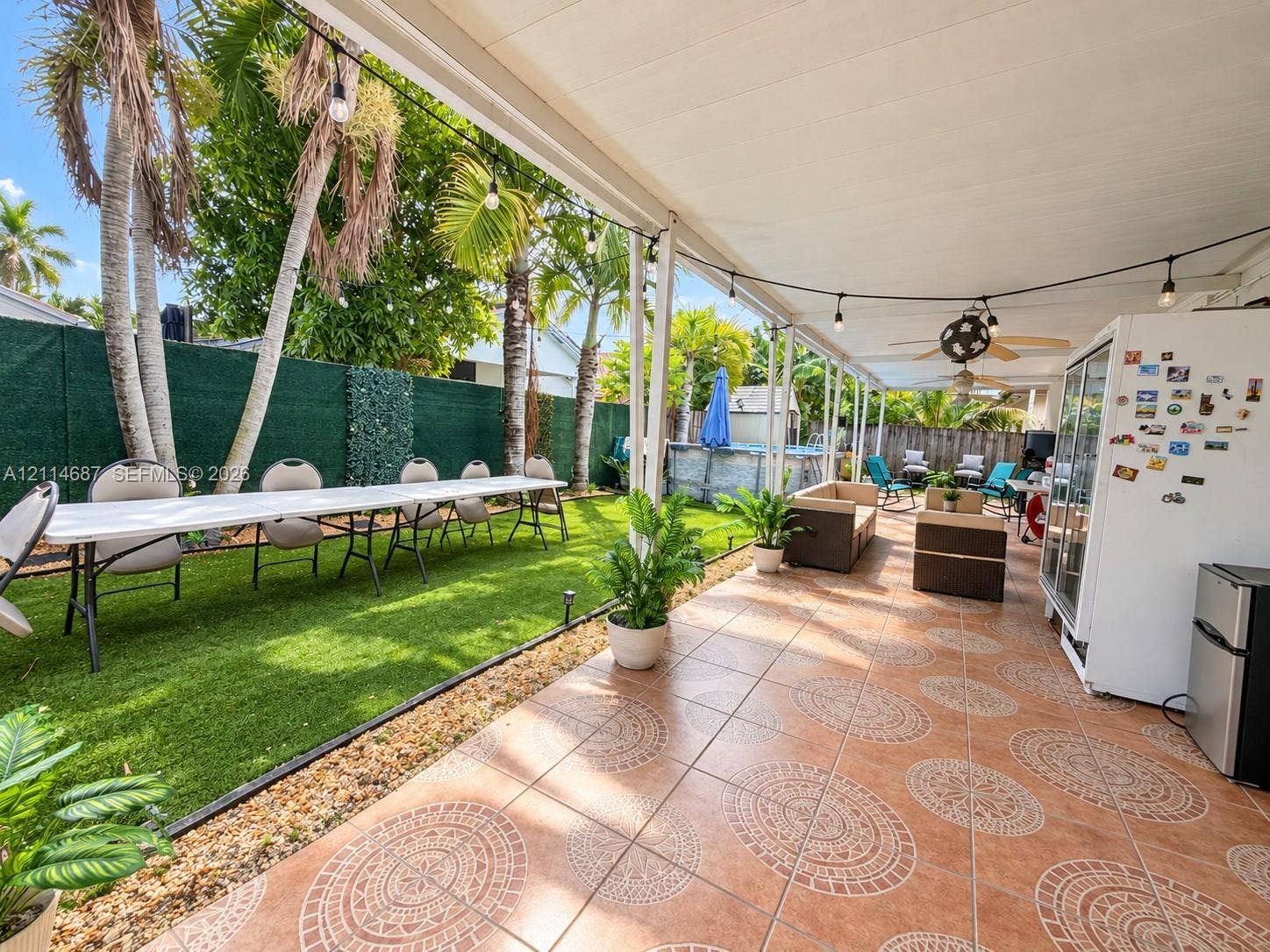 Covered patio featuring patterned tile flooring, ceiling fans, and string lighting, complemented by an artificial turf lawn and perimeter fencing