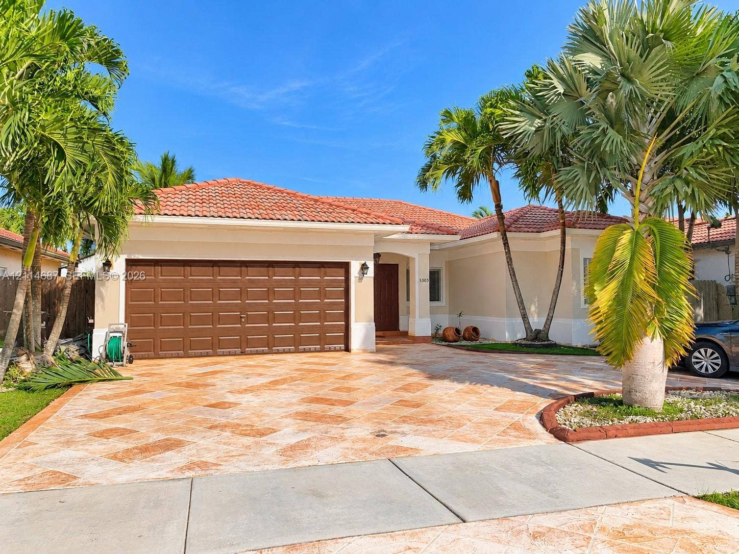 Stucco exterior featuring a terracotta tile roof, paver driveway, multi-car garage, and mature palm landscaping