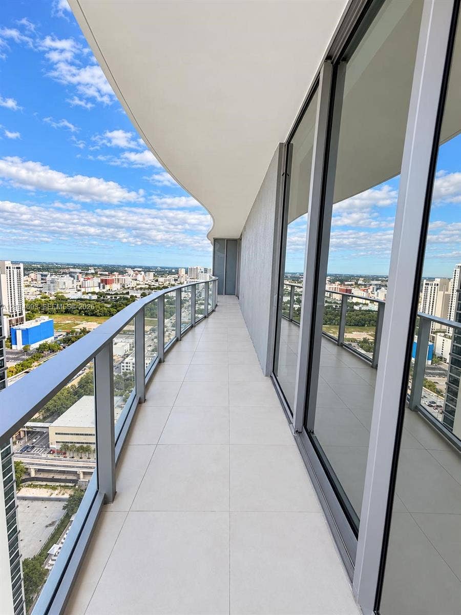 Expansive covered balcony featuring large-format tile flooring, a curved architectural ceiling, and a glass-paneled railing system with aluminum framing, bordered by floor-to-ceiling glass doors